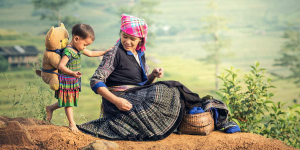 A rice farmer sitting on a rock with a small child wearing a teddybear backpack.