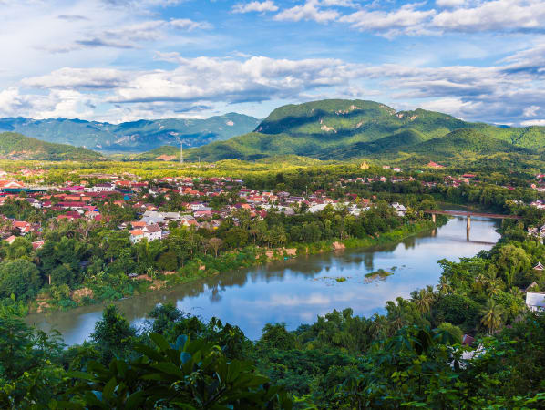 A river in Laos with rural city on its bank. Mostly white houses with red and orange roofs are nestled amongst trees and foliage. There are low mountains in the background. 