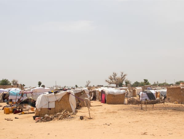 A makeshift refugee camp in a barren field. There are containers, piles of wood, and garbage strewn about.