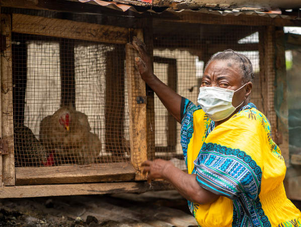 A woman in a bright yellow dress wearing a medical mask looks back at the camera, her hands are on a wooden and metal cage housing chickens. 